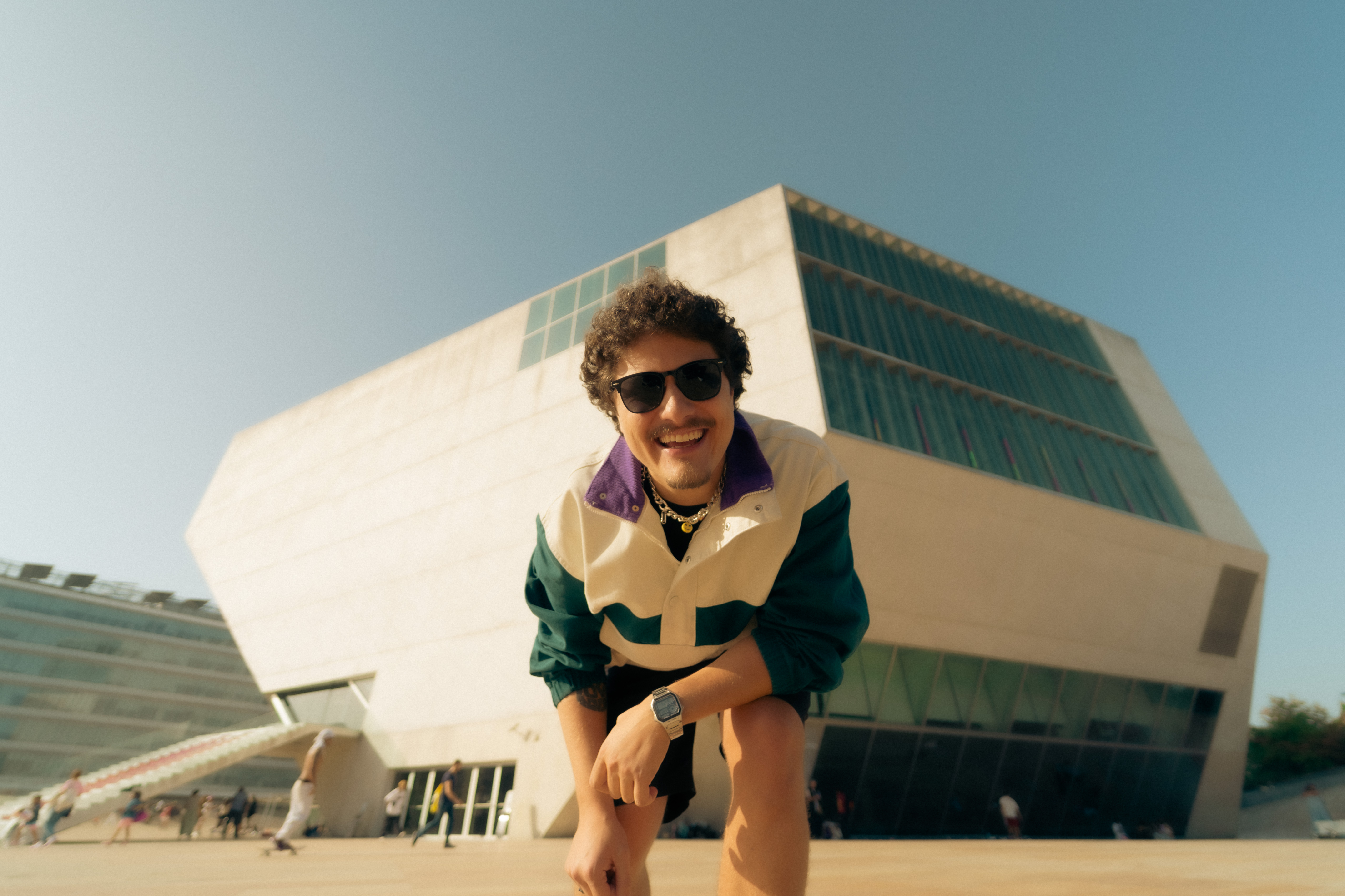 DJ Tilulu agachado e sorrindo em frente a um prédio moderno