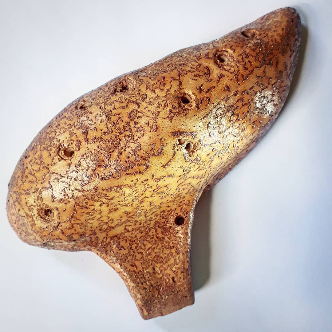 Ocarina em cerâmica de corpo alongado e curvo com base achatada, superfície ocre coberta por rede craquelada marrom-avermelhada e vários orifícios circulares de dedo
