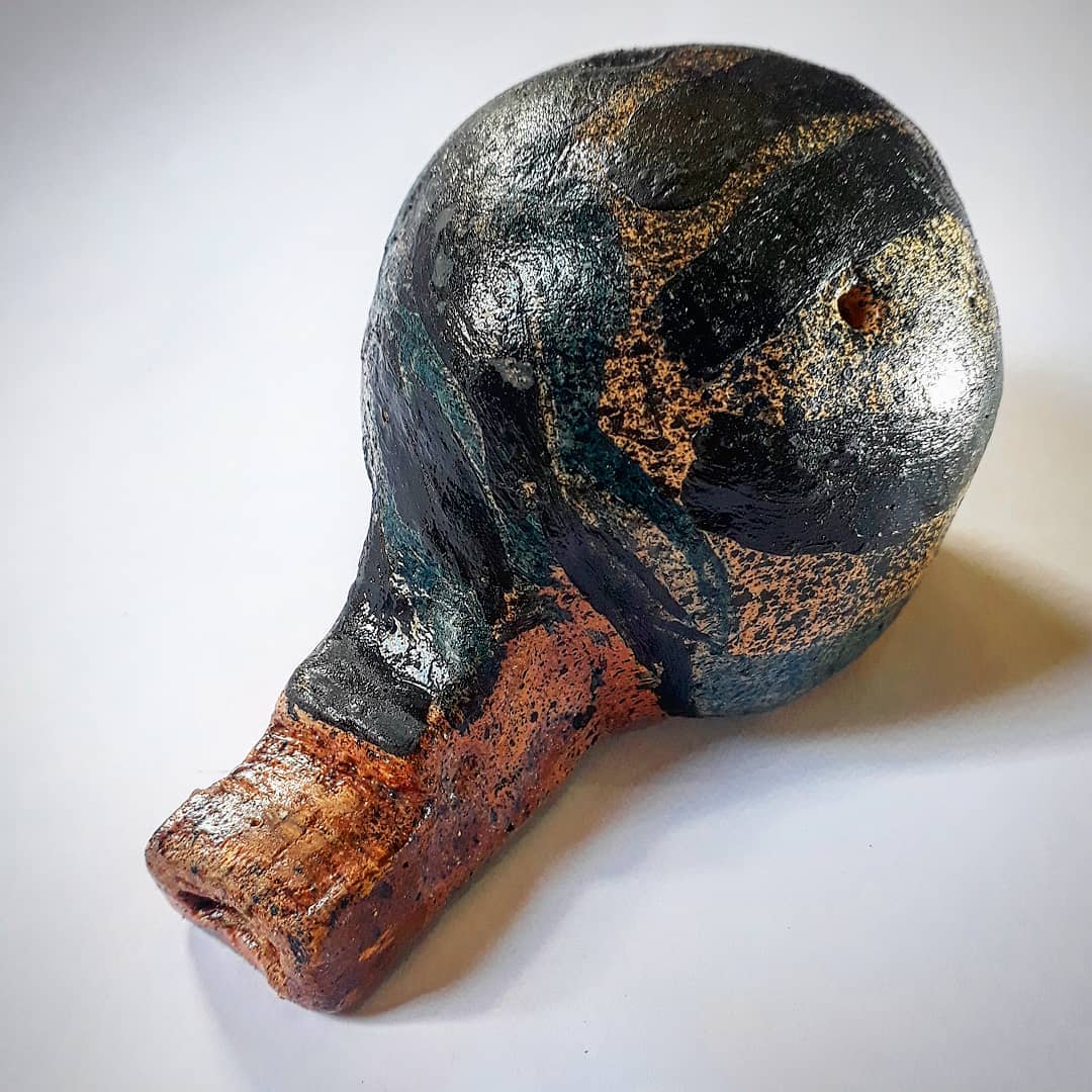 Apito em cerâmica de forma que lembra a cabeça e o bico de uma ave, com esmalte escuro brilhante salpicado de bronze metálico e bico de barro aparente