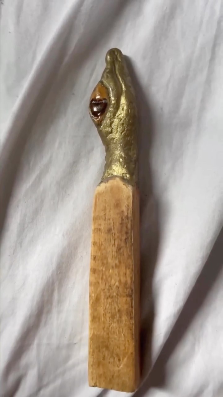 Forma orgânica de acabamento dourado texturizado, com cavidade circular escura e brilhante no topo, assentada sobre base retangular alongada de madeira de veio aparente
