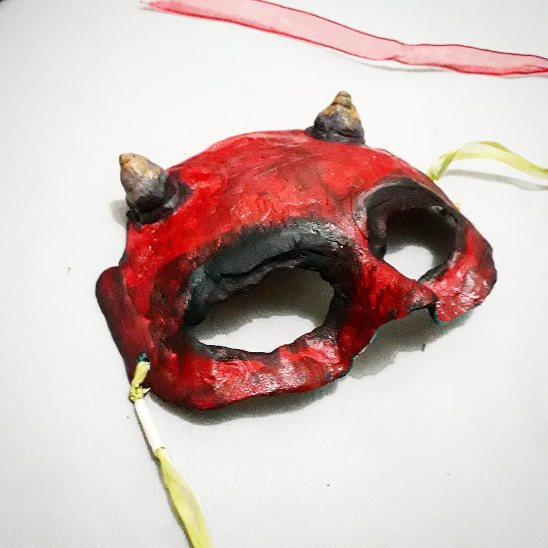 Máscara de contorno irregular em massa de papel, pintada em vermelho fosco com cavidades escuras nos olhos e na boca, duas protuberâncias cônicas no alto e cordões de tecido amarelo amarrados nas laterais