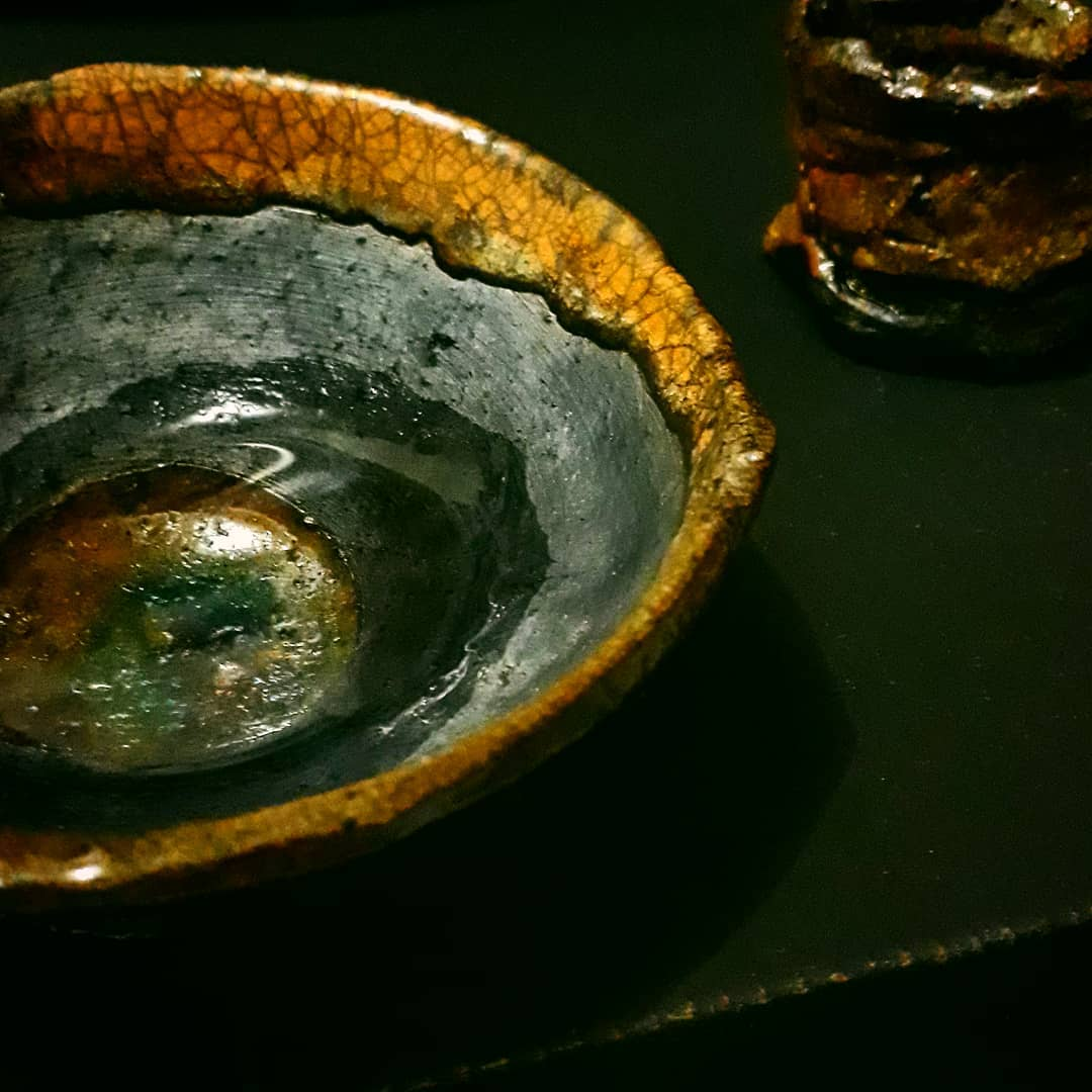 Tigela rasa de cerâmica com borda craquelada em laranja queimado e interior de esmalte escuro brilhante, com fundo iridescente verde-azulado